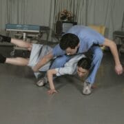 Two people in gray and blue outfits perform a complex dance or acrobatic move on a stage with hospital beds, as a third person sits in the background.