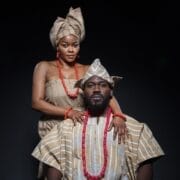 A man and woman dressed in traditional striped brown and white African attire with red bead accessories pose against a dark background, evoking regal elegance reminiscent of a Crown of Blood.