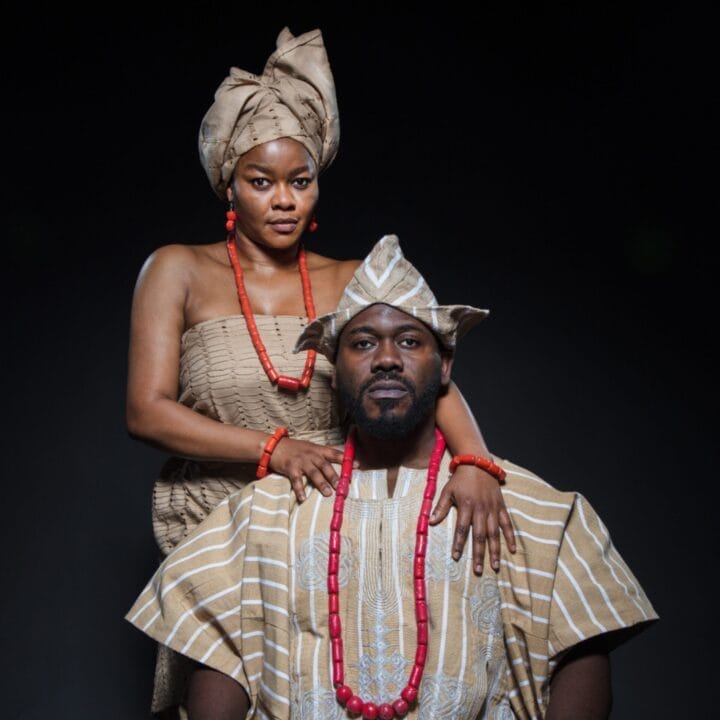 A man and woman dressed in traditional striped brown and white African attire with red bead accessories pose against a dark background, evoking regal elegance reminiscent of a Crown of Blood.