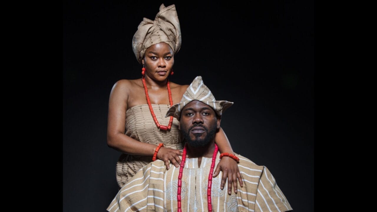 A man and woman dressed in traditional striped brown and white African attire with red bead accessories pose against a dark background, evoking regal elegance reminiscent of a Crown of Blood.