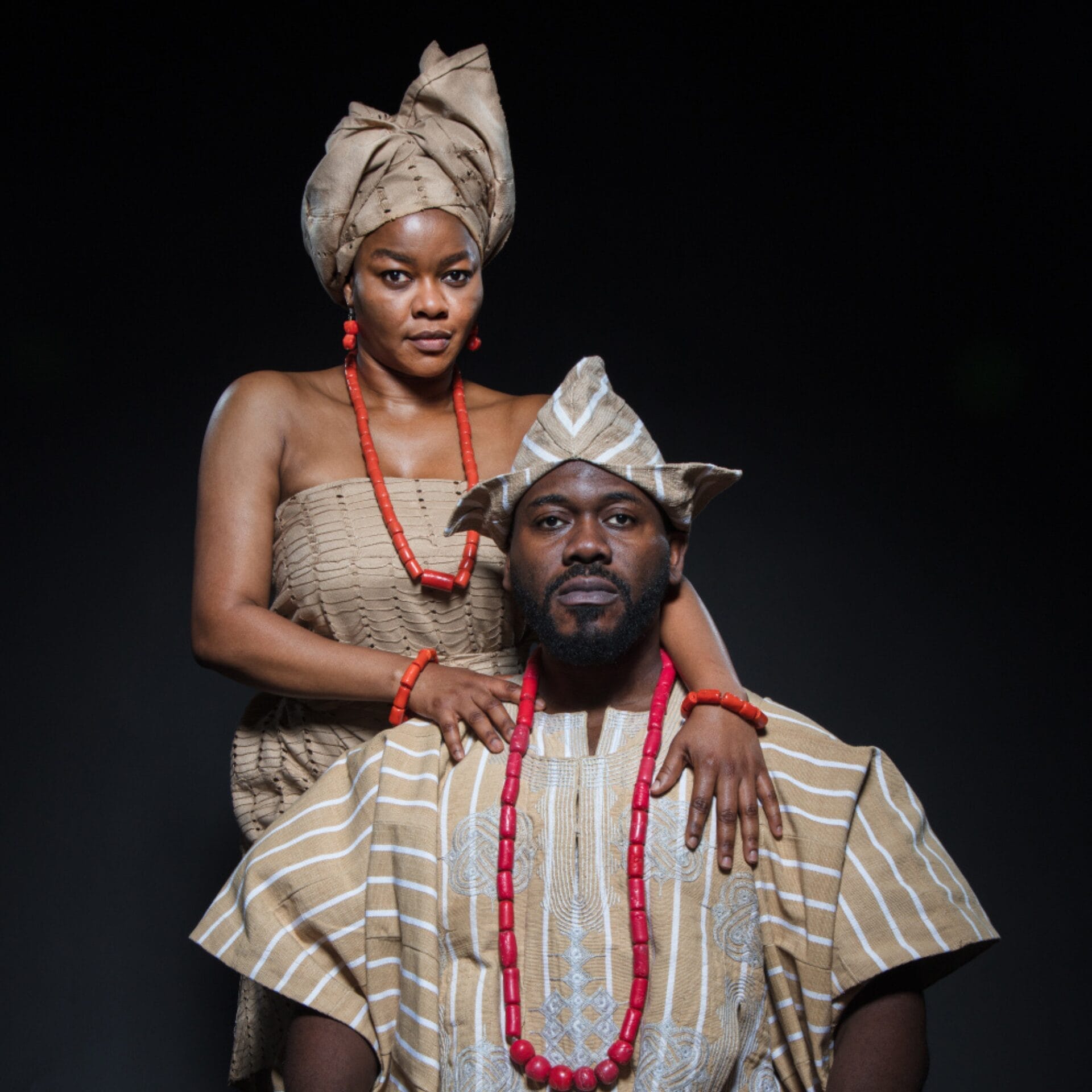 A man and woman dressed in traditional striped brown and white African attire with red bead accessories pose against a dark background, evoking regal elegance reminiscent of a Crown of Blood.