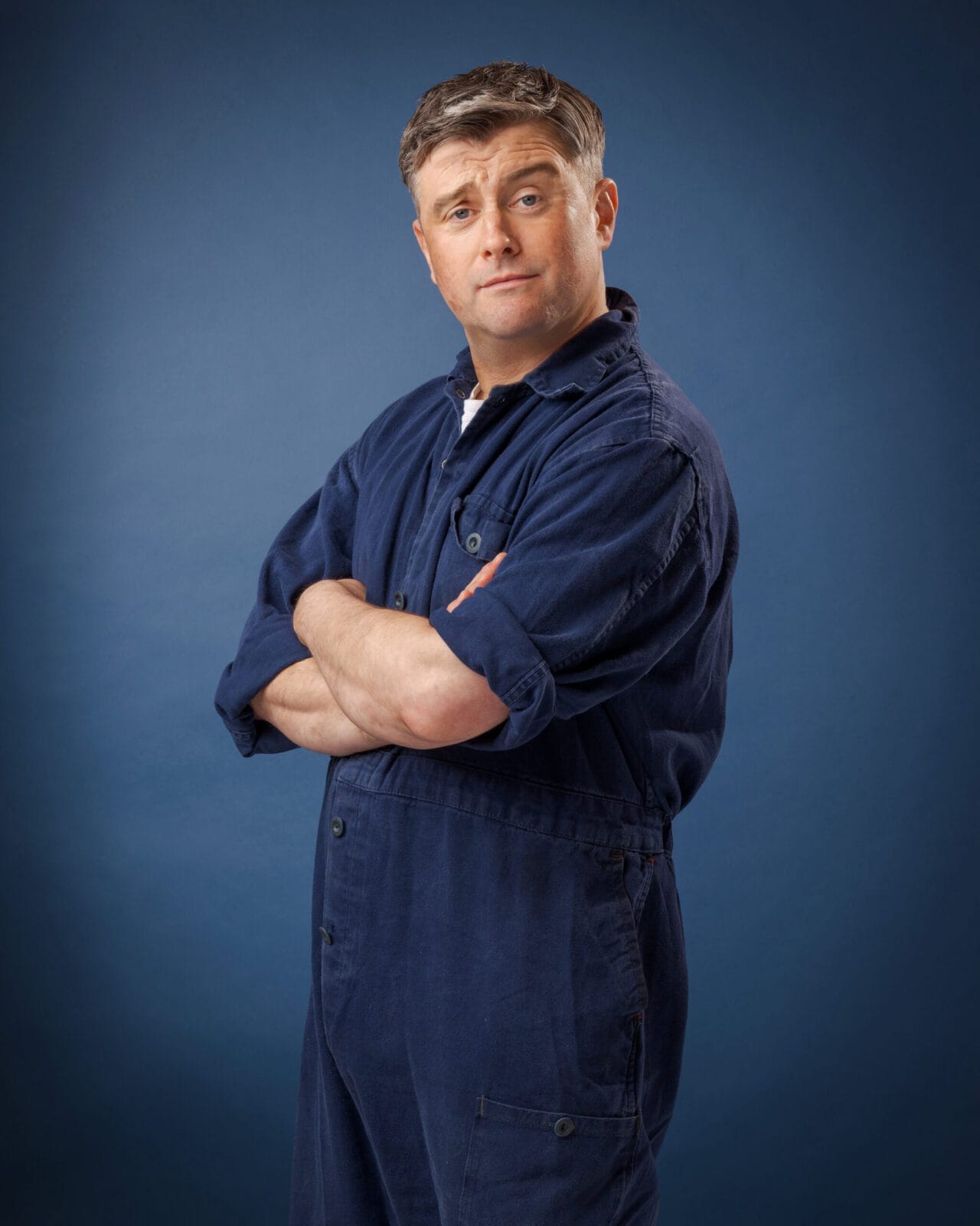 A man wearing a blue overall stands with arms crossed and a slight smirk, facing the camera against a plain blue background.