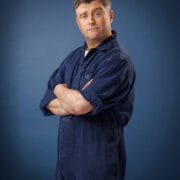 A man wearing a blue overall stands with arms crossed and a slight smirk, facing the camera against a plain blue background.