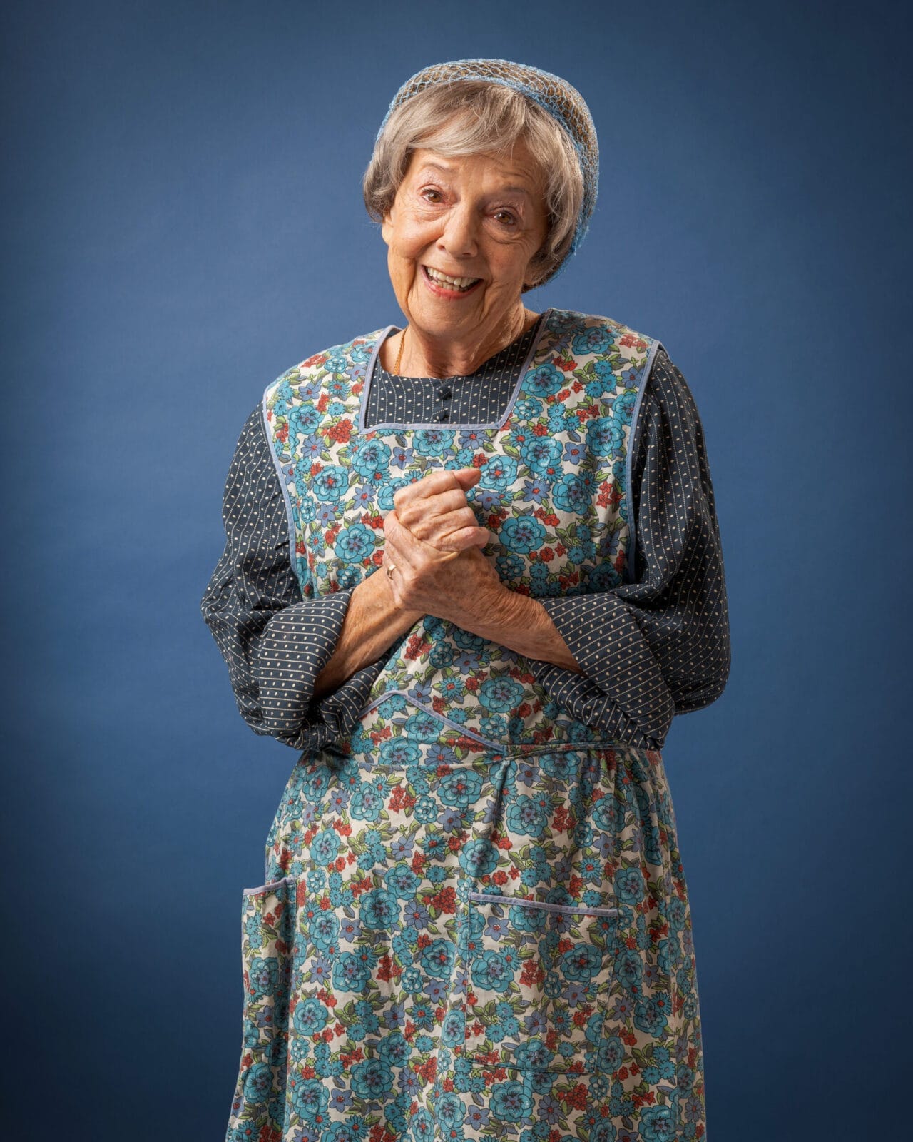 An elderly woman stands against a blue background, wearing a blue floral apron and matching headscarf over a dark, patterned long-sleeve dress. She smiles and clasps her hands in front of her chest.