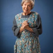 An elderly woman stands against a blue background, wearing a blue floral apron and matching headscarf over a dark, patterned long-sleeve dress. She smiles and clasps her hands in front of her chest.