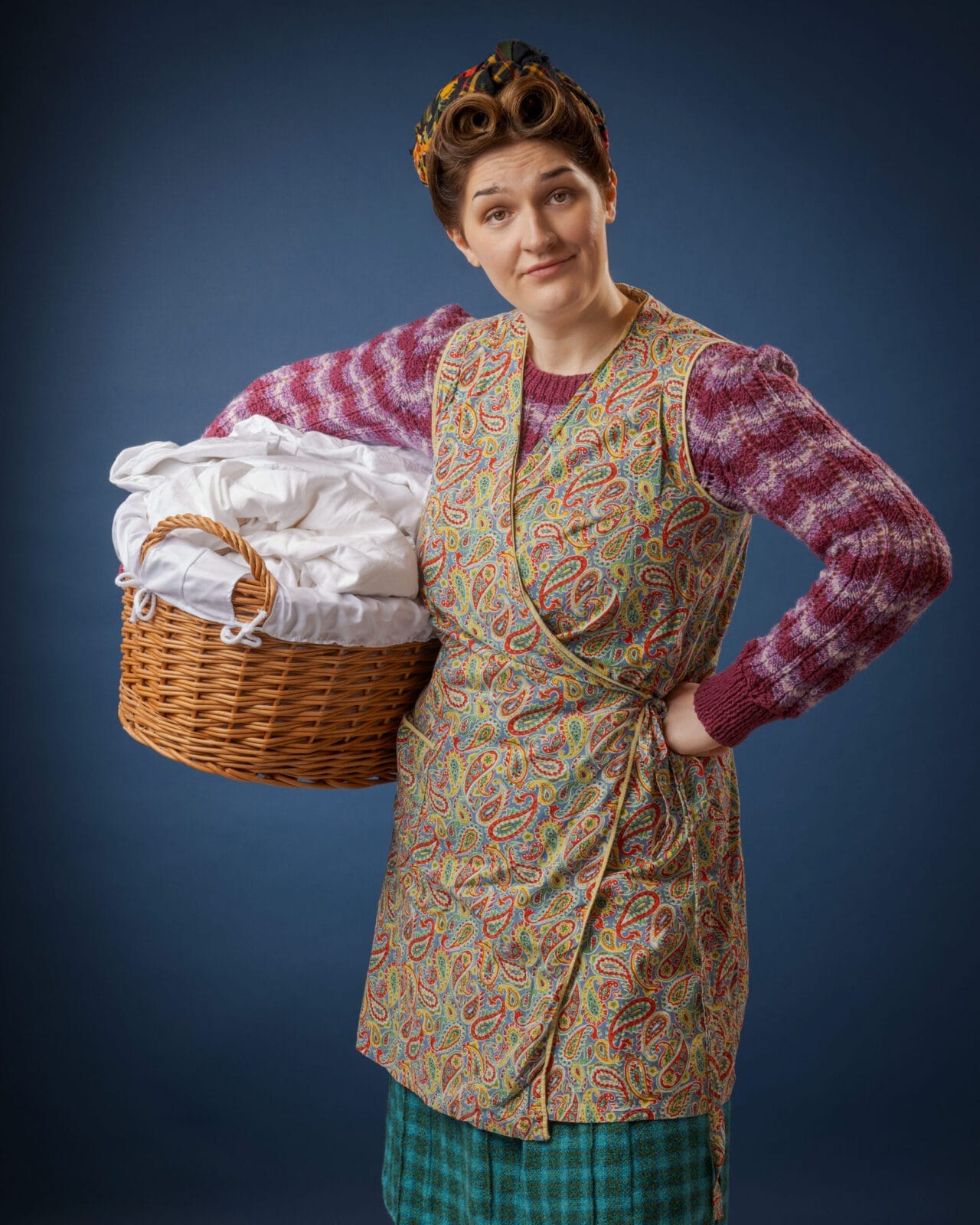 A woman with rollers in her hair holds a wicker laundry basket filled with white linen. She wears a patterned pinny over a striped jumper and checked skirt, standing against a plain blue background.