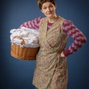 A woman with rollers in her hair holds a wicker laundry basket filled with white linen. She wears a patterned pinny over a striped jumper and checked skirt, standing against a plain blue background.