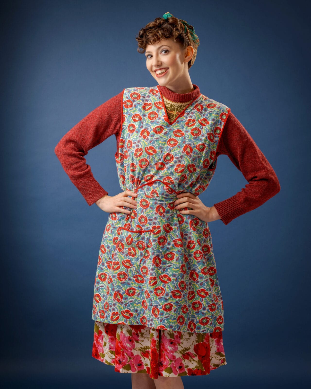 A person with short curly hair smiles, posing in a red jumper layered with a floral pinafore dress and a floral skirt. The background is plain blue, and they wear a patterned scarf tied round their head.
