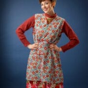 A person with short curly hair smiles, posing in a red jumper layered with a floral pinafore dress and a floral skirt. The background is plain blue, and they wear a patterned scarf tied round their head.