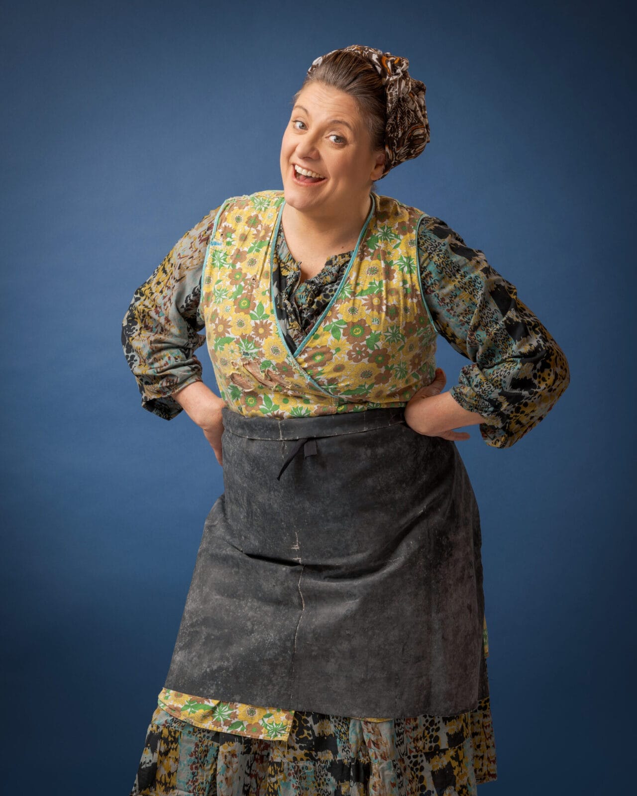 A woman wearing a patterned dress, floral apron, and headscarf stands with her hands on her hips, smiling at the camera against a plain blue background.