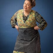 A woman wearing a patterned dress, floral apron, and headscarf stands with her hands on her hips, smiling at the camera against a plain blue background.