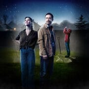 A woman and a man stand in a garden at night, echoing The Curious Incident of the Dog in the Night-Time. Behind them, a figure in a red hoodie stands by a spade and a dog-shaped chalk outline on the grass, with houses and trees looming nearby.