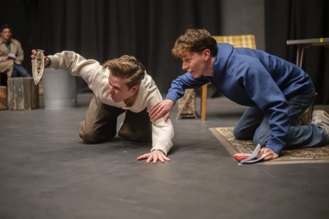 Two young men kneel on the floor of a stage. One in a white jumper holds an object out, while the other in a blue hoodie reaches towards him with one hand, holding a script in the other.