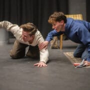 Two young men kneel on the floor of a stage. One in a white jumper holds an object out, while the other in a blue hoodie reaches towards him with one hand, holding a script in the other.
