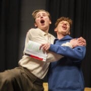 Two young men stand on stage, one holding a script with highlighted text. They are embracing each other and singing or shouting, with enthusiastic facial expressions.