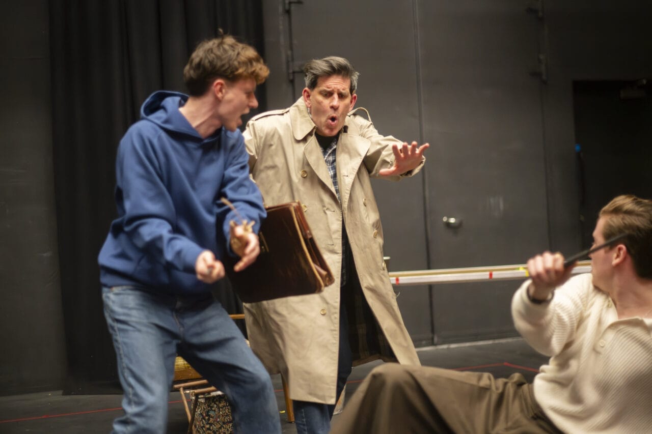 Three men act out a dramatic scene on stage. One holds a briefcase and appears startled, another in a blue hoodie gestures aggressively, and a third man sits on the floor with an arm raised defensively.