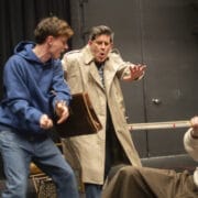 Three men act out a dramatic scene on stage. One holds a briefcase and appears startled, another in a blue hoodie gestures aggressively, and a third man sits on the floor with an arm raised defensively.