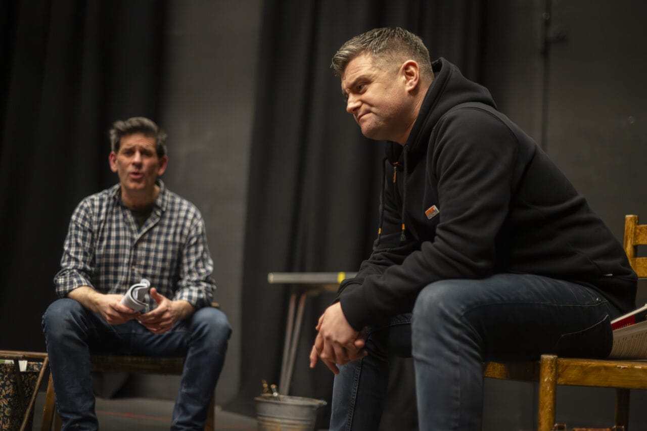 Two men are indoors on a stage. One, in a black hoodie, sits forward on a chair looking serious. The other, in a checked shirt, sits in the background appearing to speak. Black curtains and props are visible.