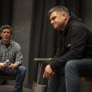 Two men are indoors on a stage. One, in a black hoodie, sits forward on a chair looking serious. The other, in a checked shirt, sits in the background appearing to speak. Black curtains and props are visible.