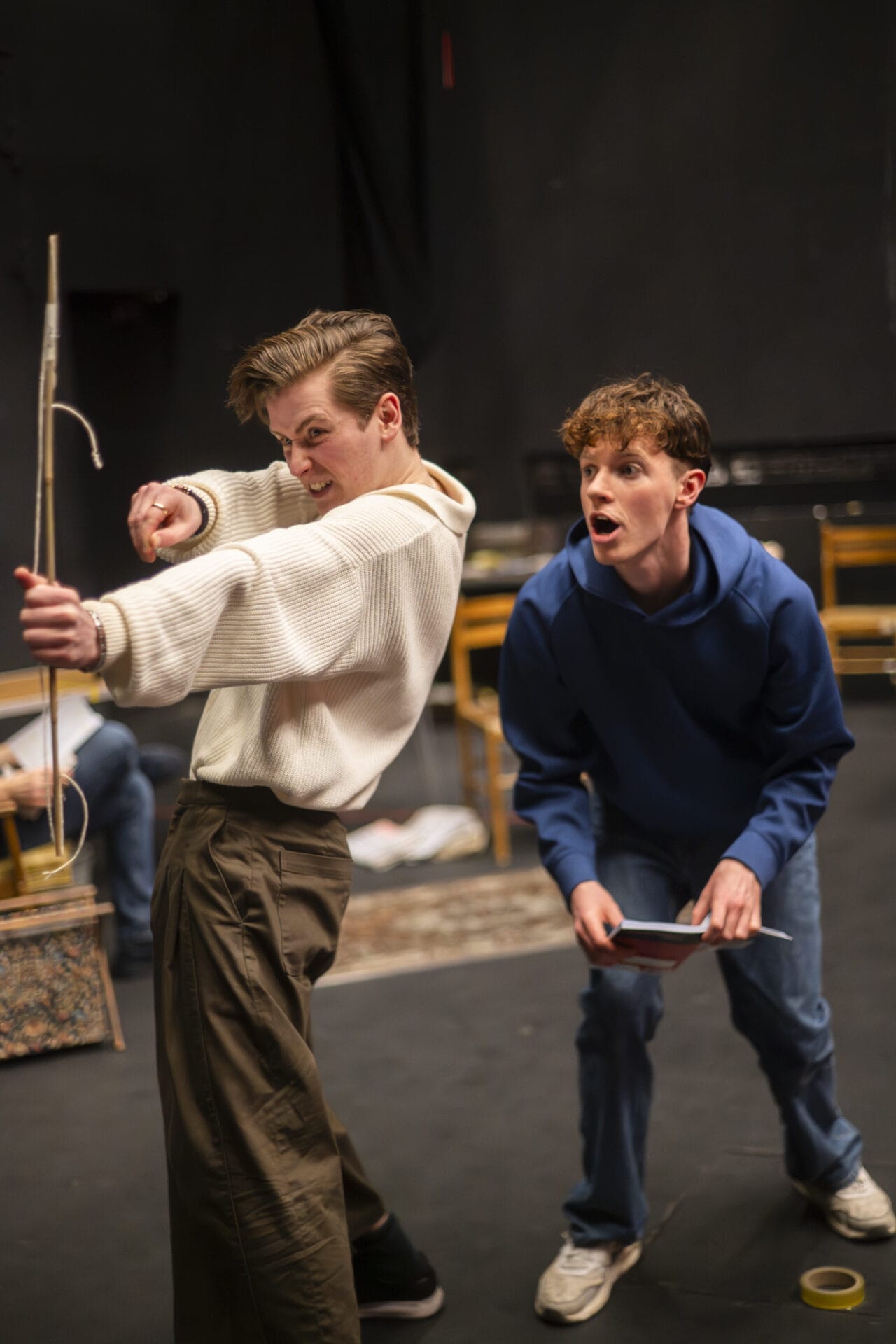 Two young men are on a stage. One is pulling back a long stick like a bow, aiming it; he wears a white jumper and brown trousers. The other, in a blue hoodie and jeans, crouches behind him, holding a small notebook and reacting.