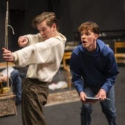 Two young men are on a stage. One is pulling back a long stick like a bow, aiming it; he wears a white jumper and brown trousers. The other, in a blue hoodie and jeans, crouches behind him, holding a small notebook and reacting.