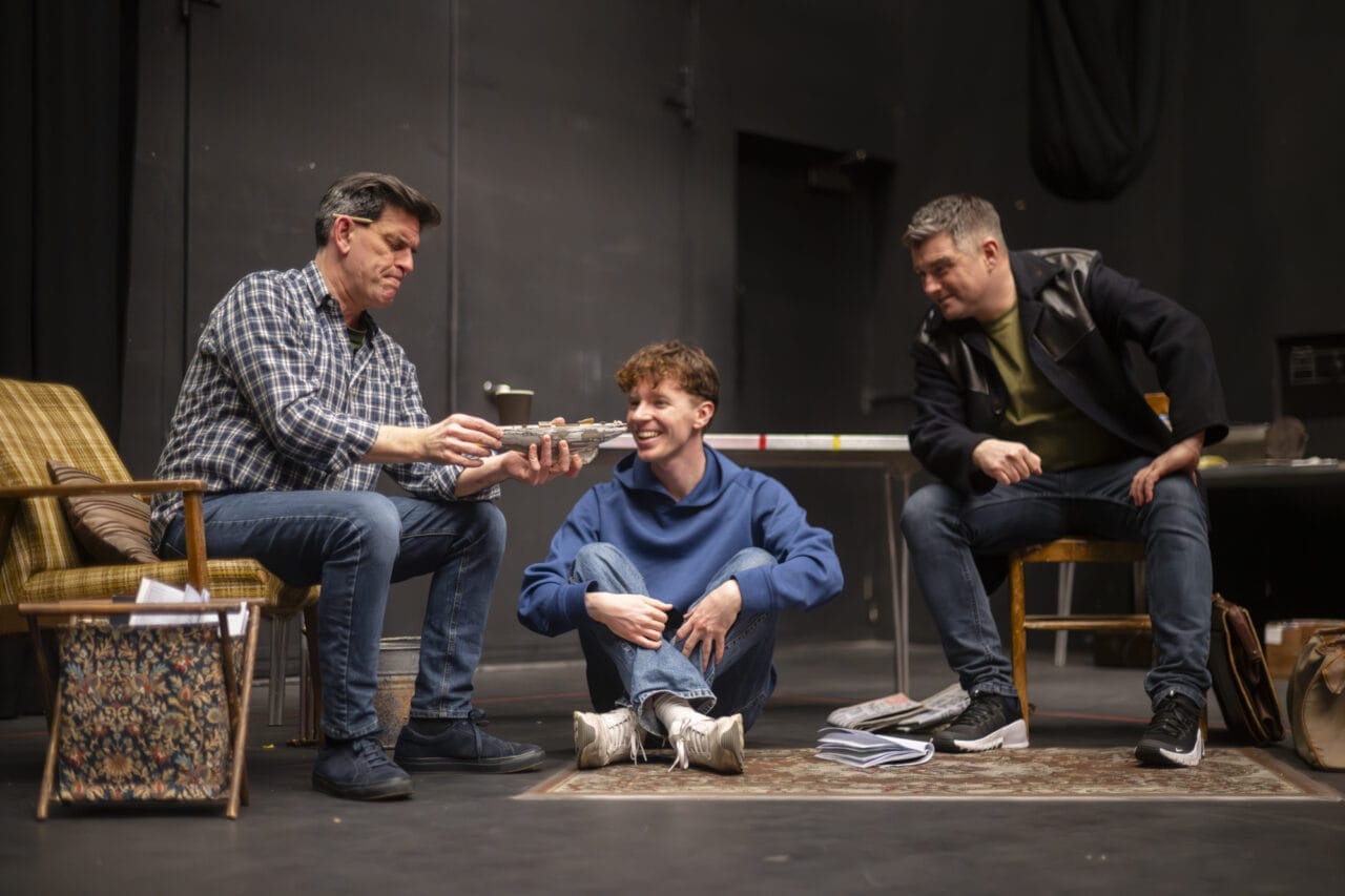 Three people interact in a room with a dark background; one sits in an armchair, one sits cross-legged on the floor smiling, and the third sits on a table. Papers and magazines are scattered on the floor.