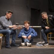 Three people interact in a room with a dark background; one sits in an armchair, one sits cross-legged on the floor smiling, and the third sits on a table. Papers and magazines are scattered on the floor.