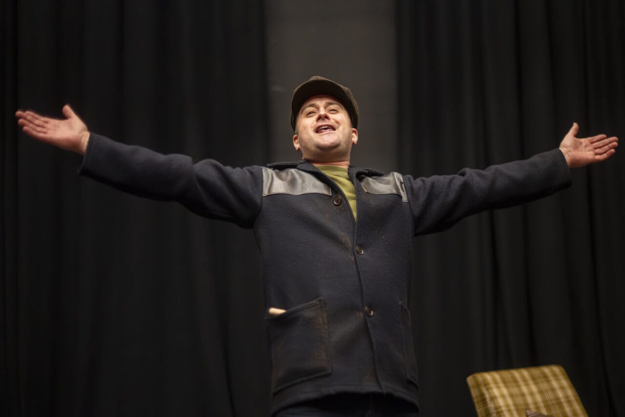 A person wearing a dark coat and a cap stands with arms outstretched, facing forwards.