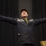 A person wearing a dark coat and a cap stands with arms outstretched, facing forwards.