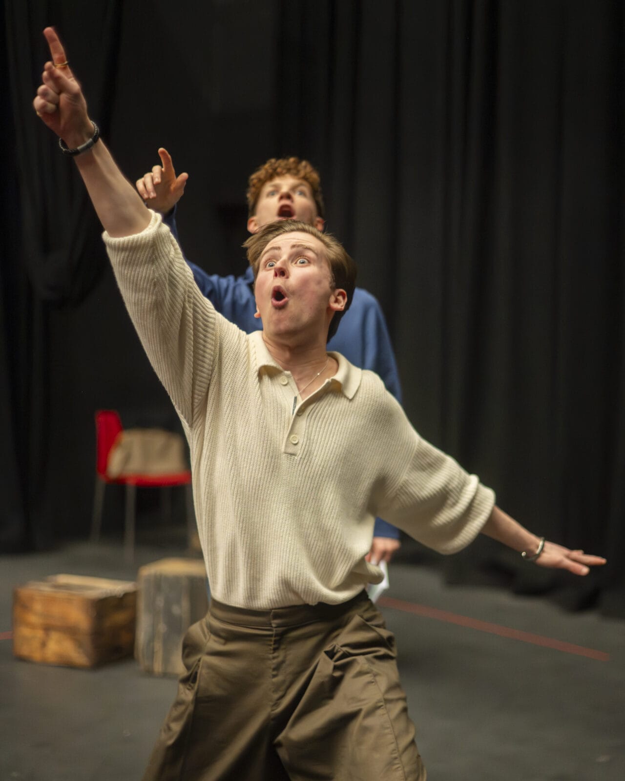 Two people perform on stage; the person in front gestures upward with an expressive face, whilst the person behind also points up.