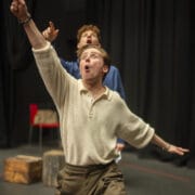 Two people perform on stage; the person in front gestures upward with an expressive face, whilst the person behind also points up.