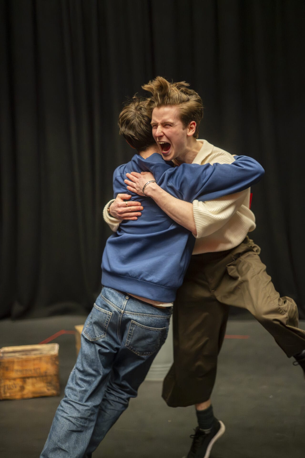 Two people hug on a stage with dark curtains behind them. One person, wearing a light jumper and dark trousers, appears to be shouting or expressing excitement. The other person wears a blue jacket and jeans.
