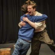 Two people hug on a stage with dark curtains behind them. One person, wearing a light jumper and dark trousers, appears to be shouting or expressing excitement. The other person wears a blue jacket and jeans.