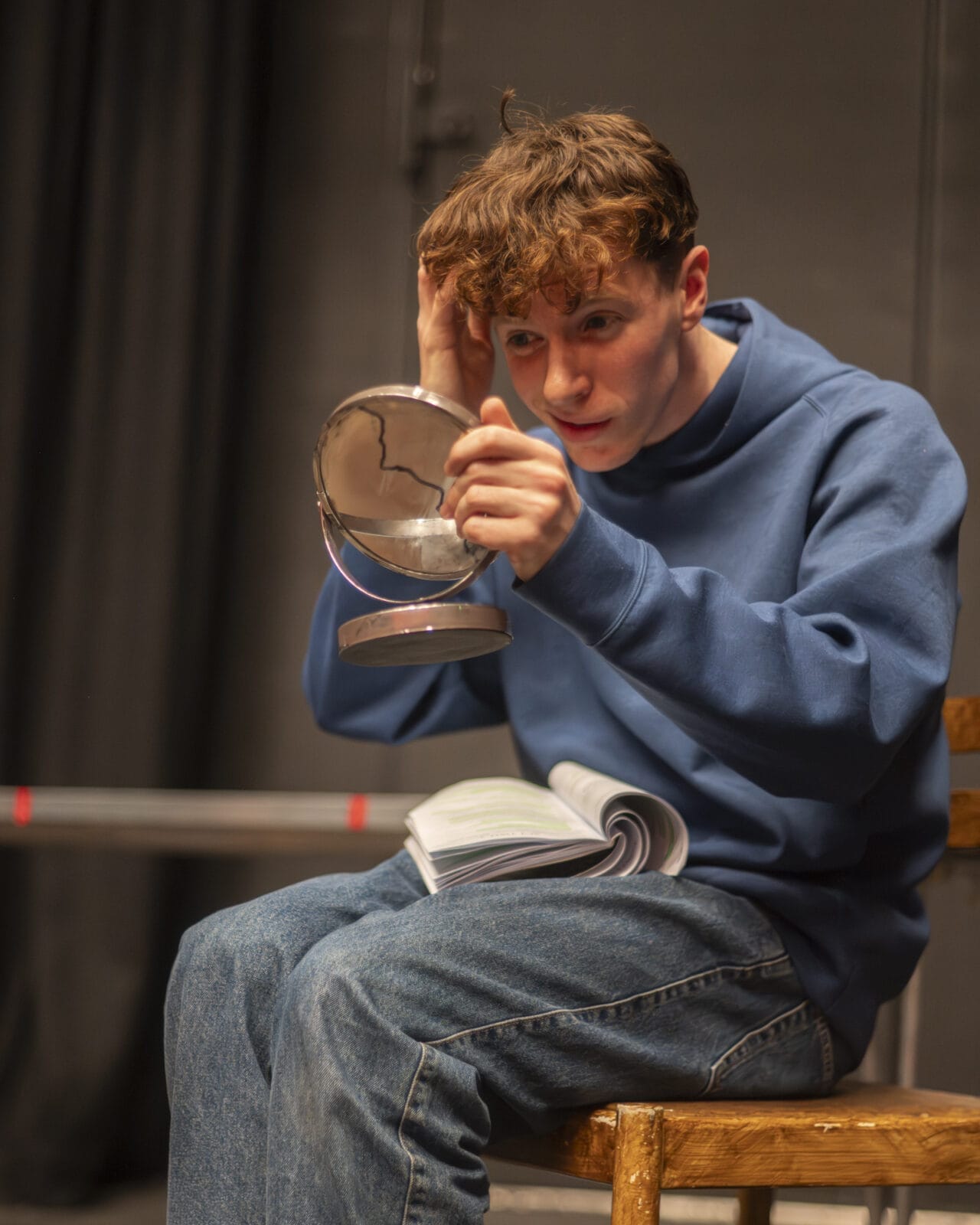 A young person in a blue hoodie and jeans sits on a wooden chair, holding a cracked round mirror and touching their hair whilst looking into it. An open book rests on their lap. The background is out of focus.