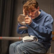 A young person in a blue hoodie and jeans sits on a wooden chair, holding a cracked round mirror and touching their hair whilst looking into it. An open book rests on their lap. The background is out of focus.