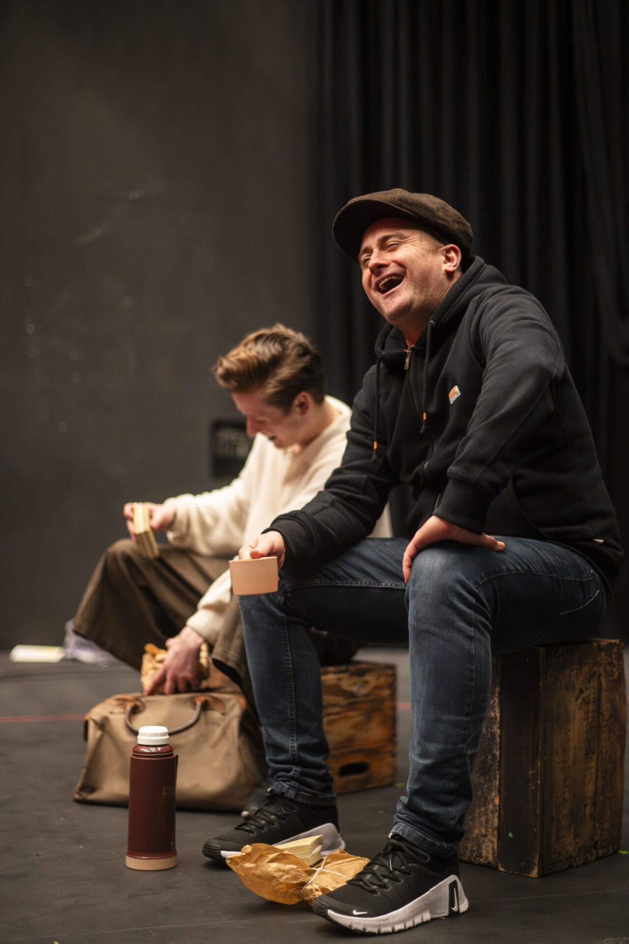 Two people sit on wooden boxes on a stage. One, in a black hoodie and cap, laughs while holding a cup. The other, in a cream jumper, looks down while reaching into a bag.