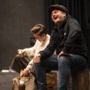 Two people sit on wooden boxes on a stage. One, in a black hoodie and cap, laughs while holding a cup. The other, in a cream jumper, looks down while reaching into a bag.