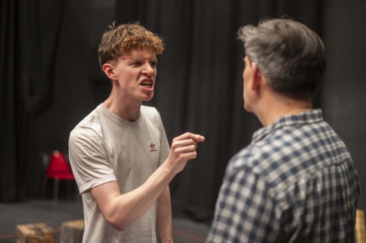 A young man in a white T-shirt points and speaks angrily to another man in a chequered shirt, who stands with his back to the camera.