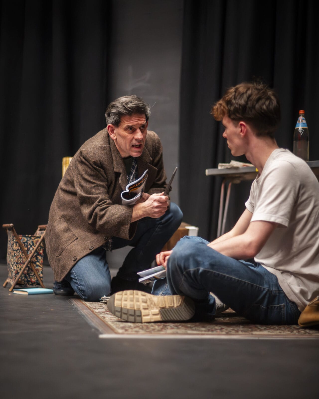 A man in a brown coat kneels on a rug holding scissors, speaking intensely to a seated young man in a white T-shirt.