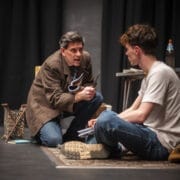 A man in a brown coat kneels on a rug holding scissors, speaking intensely to a seated young man in a white T-shirt.