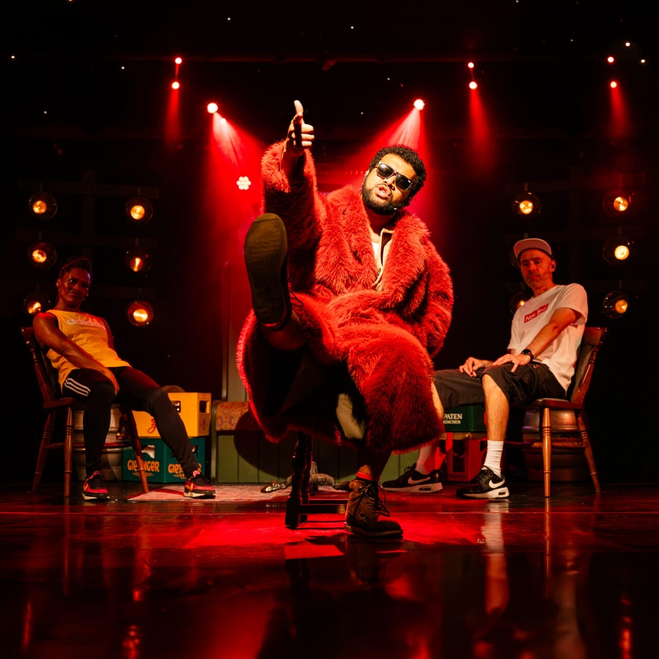 A person in a red fur coat sits on a chair with one leg raised and gives a thumbs-up. Two others sit on either side, one in a yellow shirt and the other in white, against a backdrop of red stage lights.