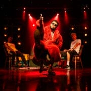 A person in a red fur coat sits on a chair with one leg raised and gives a thumbs-up. Two others sit on either side, one in a yellow shirt and the other in white, against a backdrop of red stage lights.