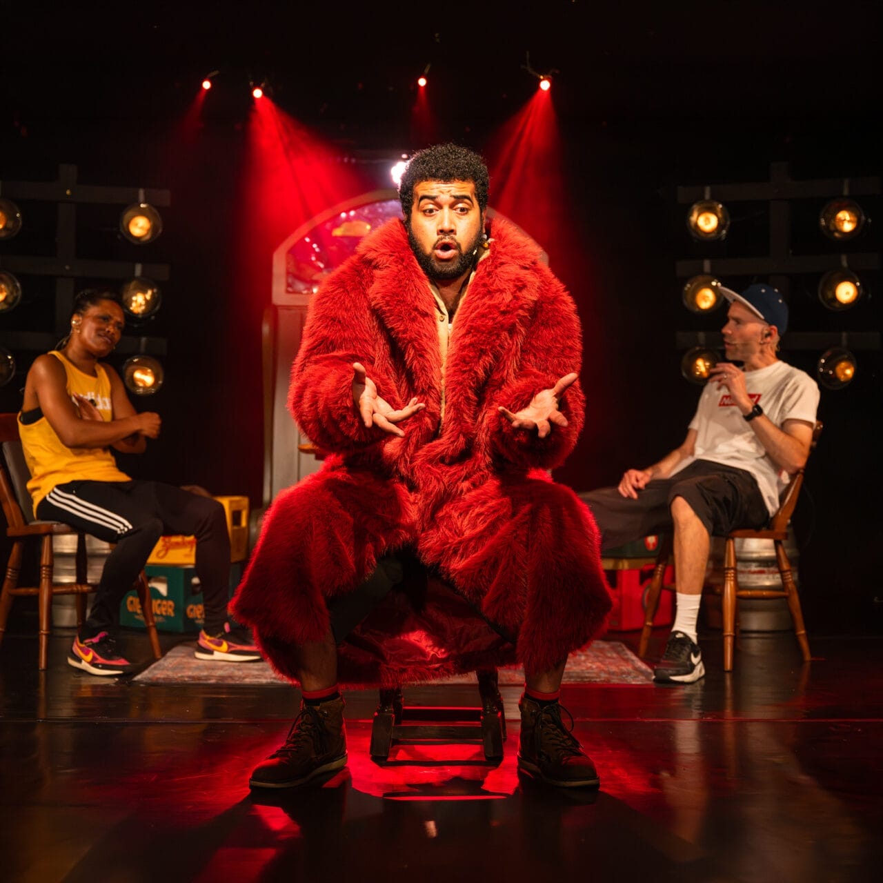 A man in a large red fur coat sits on a stool centre stage under dramatic red lighting, gesturing with his hands. Two other people sit in the background, one wearing a yellow vest and the other a white t-shirt and cap.