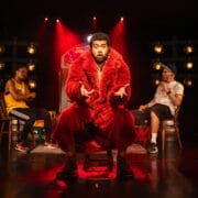 A man in a large red fur coat sits on a stool centre stage under dramatic red lighting, gesturing with his hands. Two other people sit in the background, one wearing a yellow vest and the other a white t-shirt and cap.