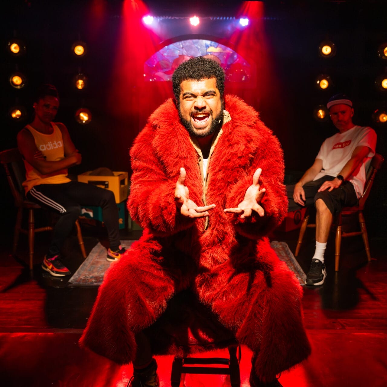 A man in a red fur coat sits centre stage under dramatic red lighting, gesturing with both hands. Two people sit behind him, one on each side, with a dark background and multiple lights visible.