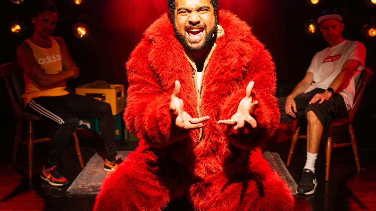 A man in a red fur coat sits centre stage under dramatic red lighting, gesturing with both hands. Two people sit behind him, one on each side, with a dark background and multiple lights visible.