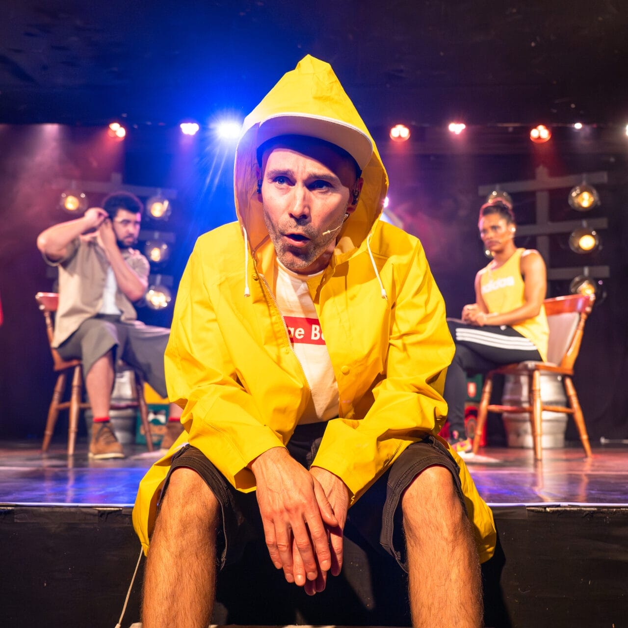 A man in a yellow raincoat and hat sits on stage under bright lights, looking intently forward. Two people sit behind him on chairs, with stage lighting and props in the background.