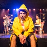 A man in a yellow raincoat and hat sits on stage under bright lights, looking intently forward. Two people sit behind him on chairs, with stage lighting and props in the background.