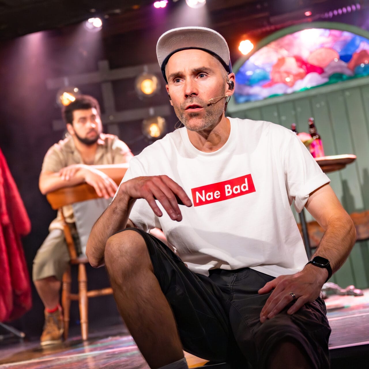 A man wearing a headset, white "Nae Bad" t-shirt, shorts, and a cap sits onstage in the foreground. Another man sits at a table in the background. Stage lighting and set pieces are visible.