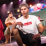 A man wearing a headset, white "Nae Bad" t-shirt, shorts, and a cap sits onstage in the foreground. Another man sits at a table in the background. Stage lighting and set pieces are visible.